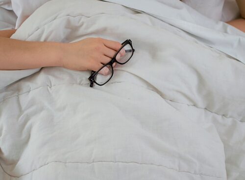 person lying on bed while covering face with pillow and holding eyeglasses