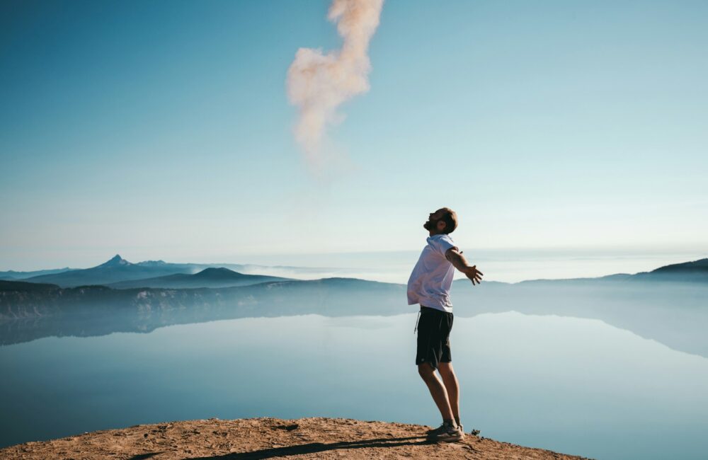 man standing on sand while spreading arms beside calm body of water