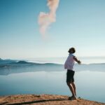 man standing on sand while spreading arms beside calm body of water
